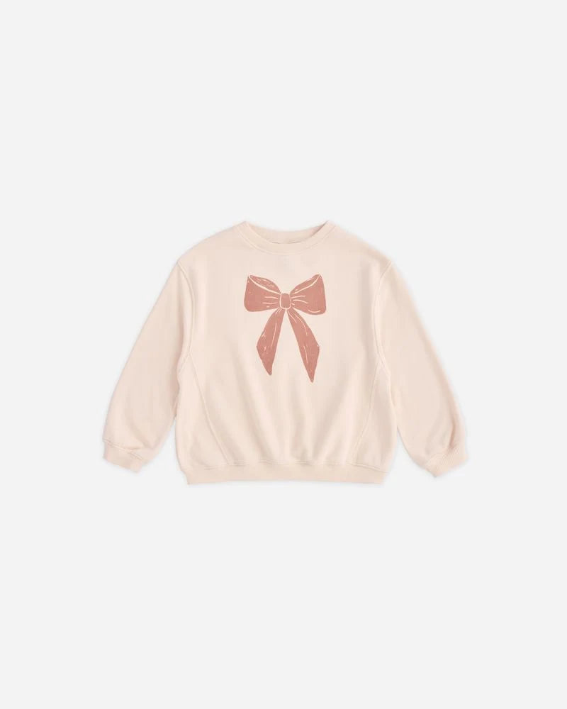 A light peach crewneck sweatshirt featuring a textured dusty rose bow graphic printed on the chest, displayed flat against a clean off-white background.